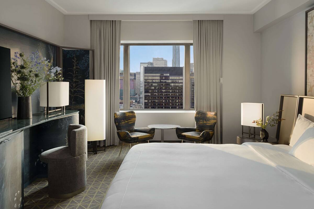 A hotel room with a large bed, two armchairs, a small table, a desk with a TV, and a large window offering a city view.