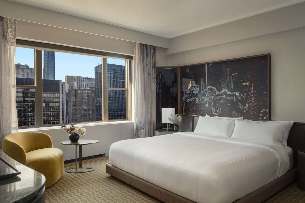 A hotel room with a large bed, a small round table, a yellow armchair, and a view of city buildings through large windows.