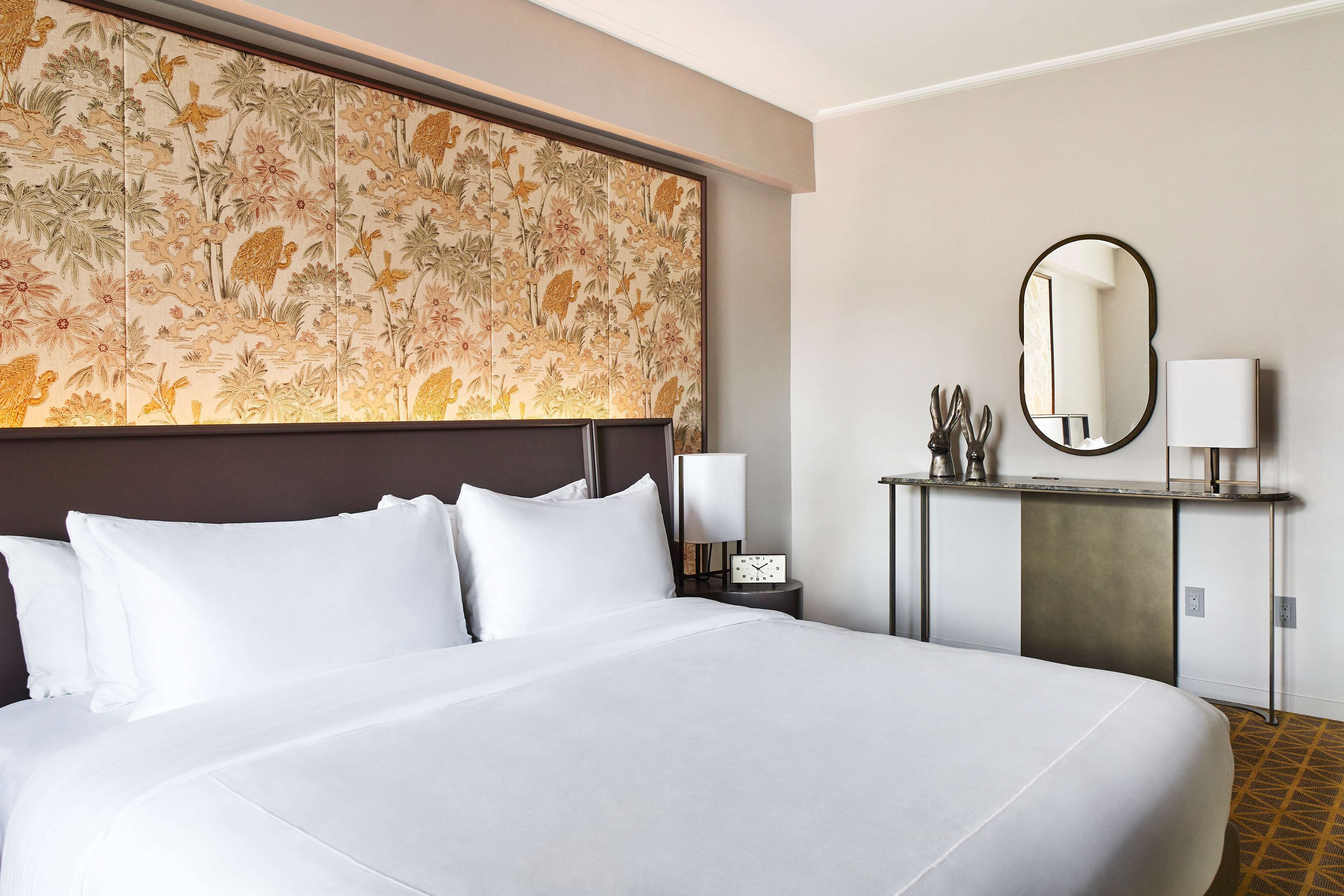 A hotel room with a large bed, a headboard with a floral design, a bedside table with a lamp, a mirror, and a small cabinet.