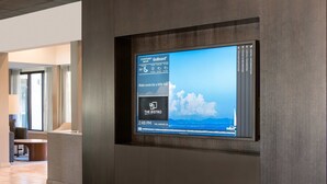 Un écran numérique dans le hall d’un hôtel, offrant une vue sur la mer et les montagnes, une horloge indiquant 14 h 48, et une enseigne indiquant « The Bistro ».