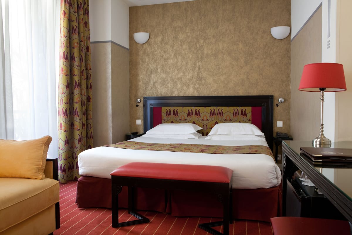 Ein Hotelzimmer mit einem großen Bett, einer roten Bank, einem Schreibtisch mit Lampe und einem Sofa.