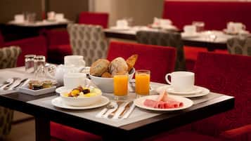 A breakfast setting with a table set for two, including plates of food, a bowl of fruit, two glasses of orange juice, and a variety of pastries.