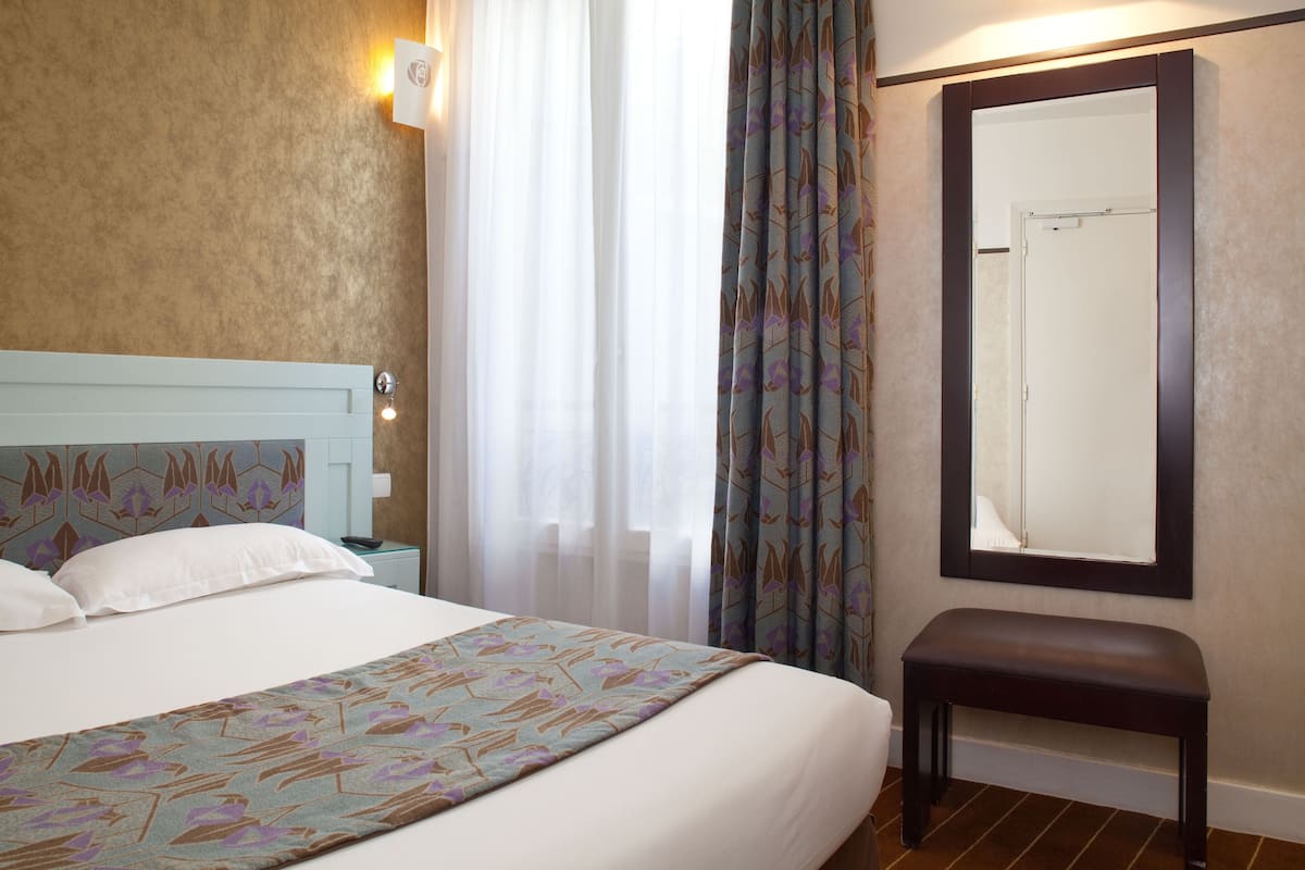 Ein Hotelzimmer mit einem Bett, einem Nachttisch, einer Bank, einem Spiegel und einem Fenster mit Vorhängen.