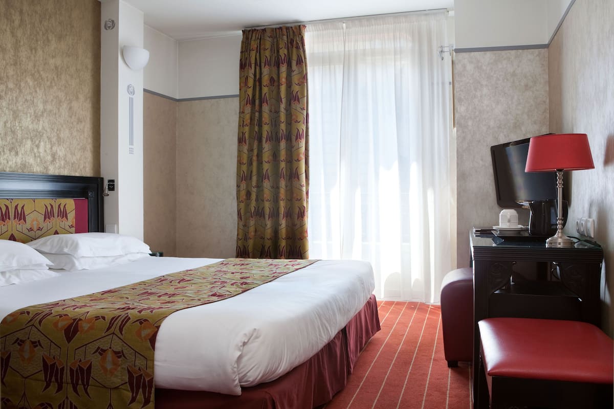 Ein Hotelzimmer mit einem Bett, einem Schreibtisch mit Lampe, einem Sessel und einem Fenster mit Vorhängen.