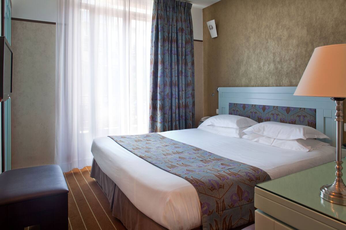 Ein Hotelzimmer mit einem Bett, einem Nachttisch, einer Lampe, einer Bank und einem Fenster mit Vorhängen.