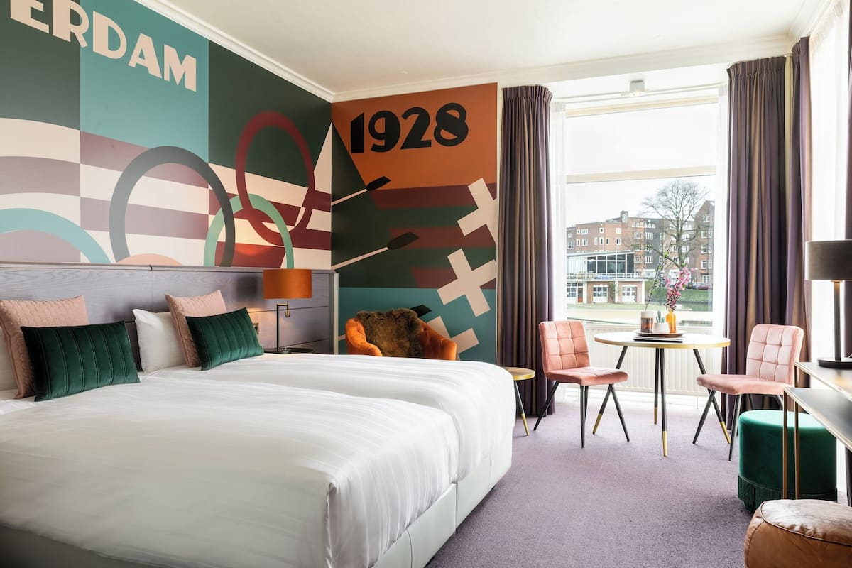 A hotel room with a large bed, two chairs, a small table, and a view of the city through large windows.