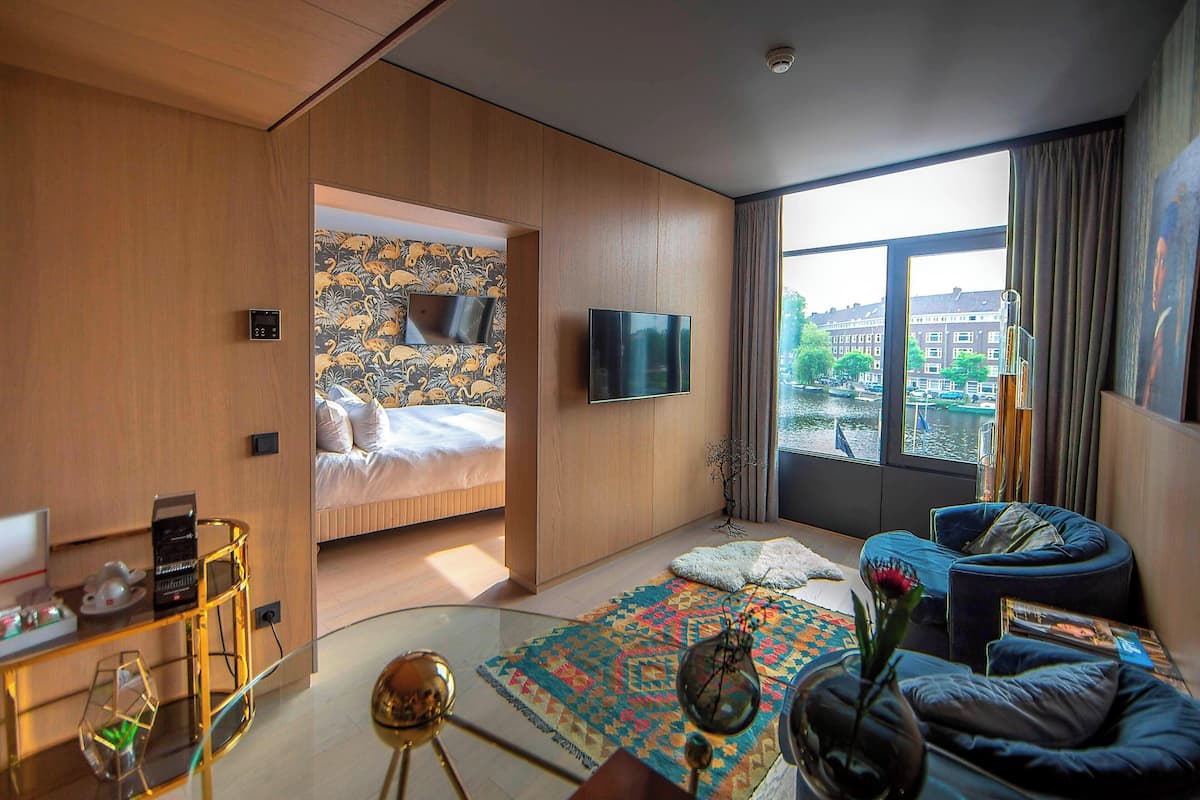A modern hotel room with a bed, a TV, a desk with a computer, and a view of the city through large windows.