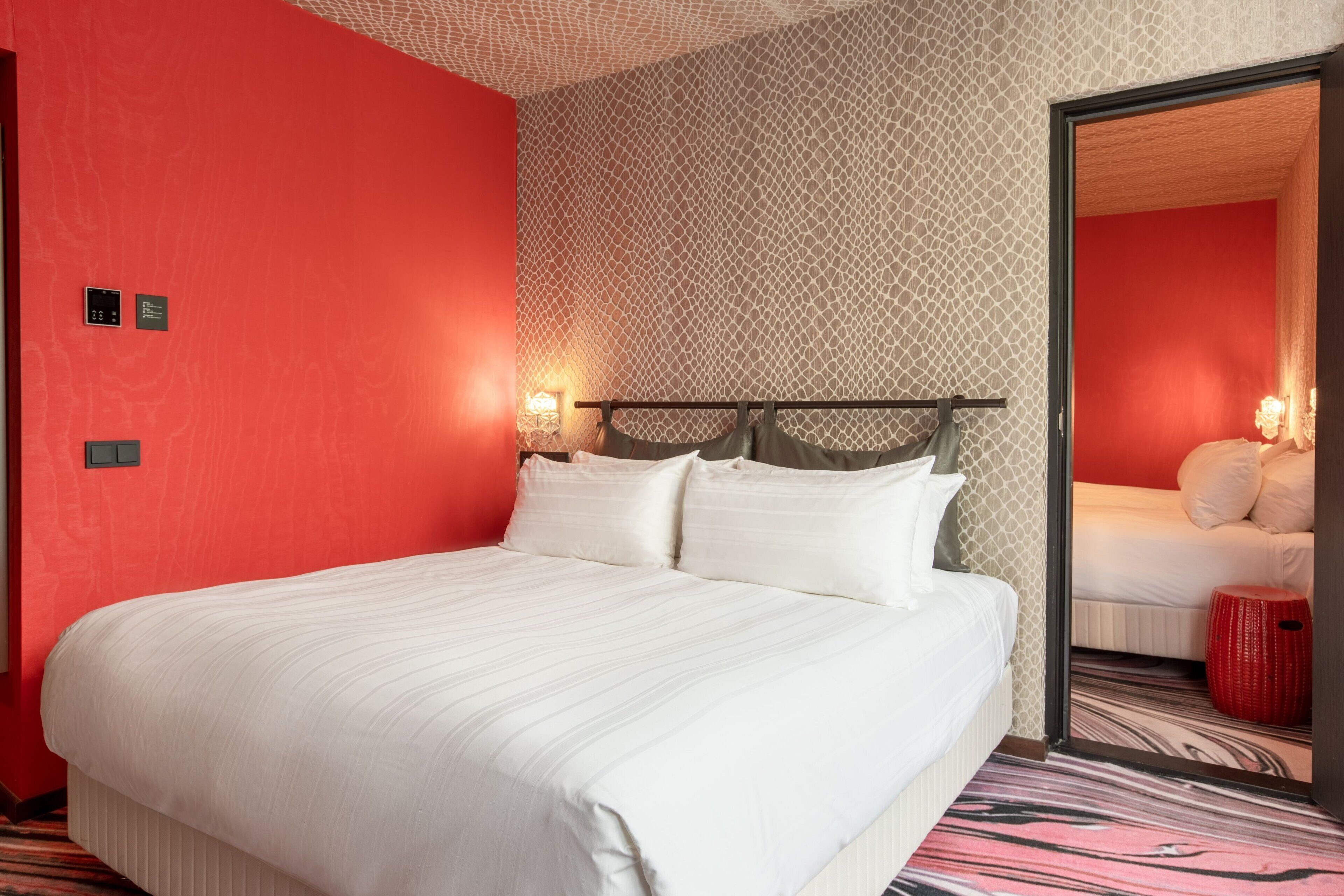 A hotel room with a large bed, a red accent wall, a patterned wallpaper, and a mirror reflecting another bed.