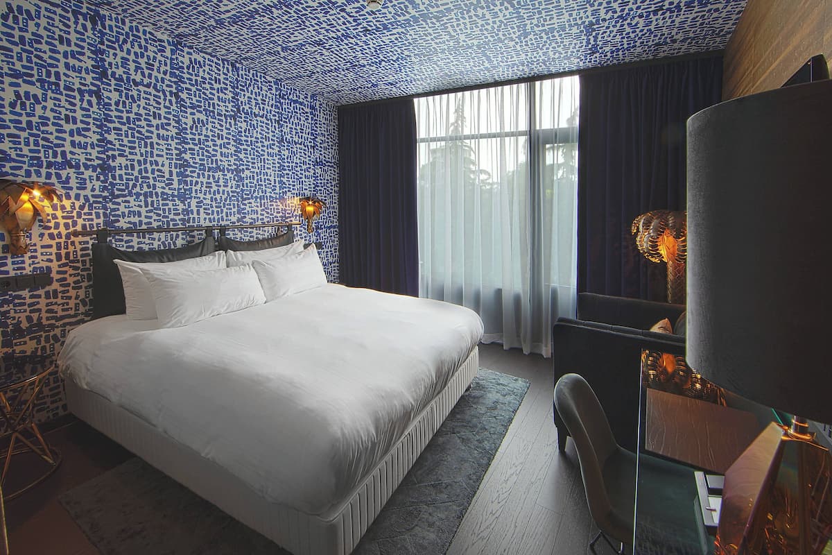 A hotel room with a large bed, a desk, and a chair. There is a window with curtains, and the walls are covered in a blue mosaic pattern.