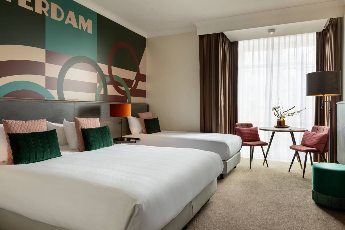 A hotel room with two beds, a small table with chairs, a lamp, and a view of the outdoors through large windows.