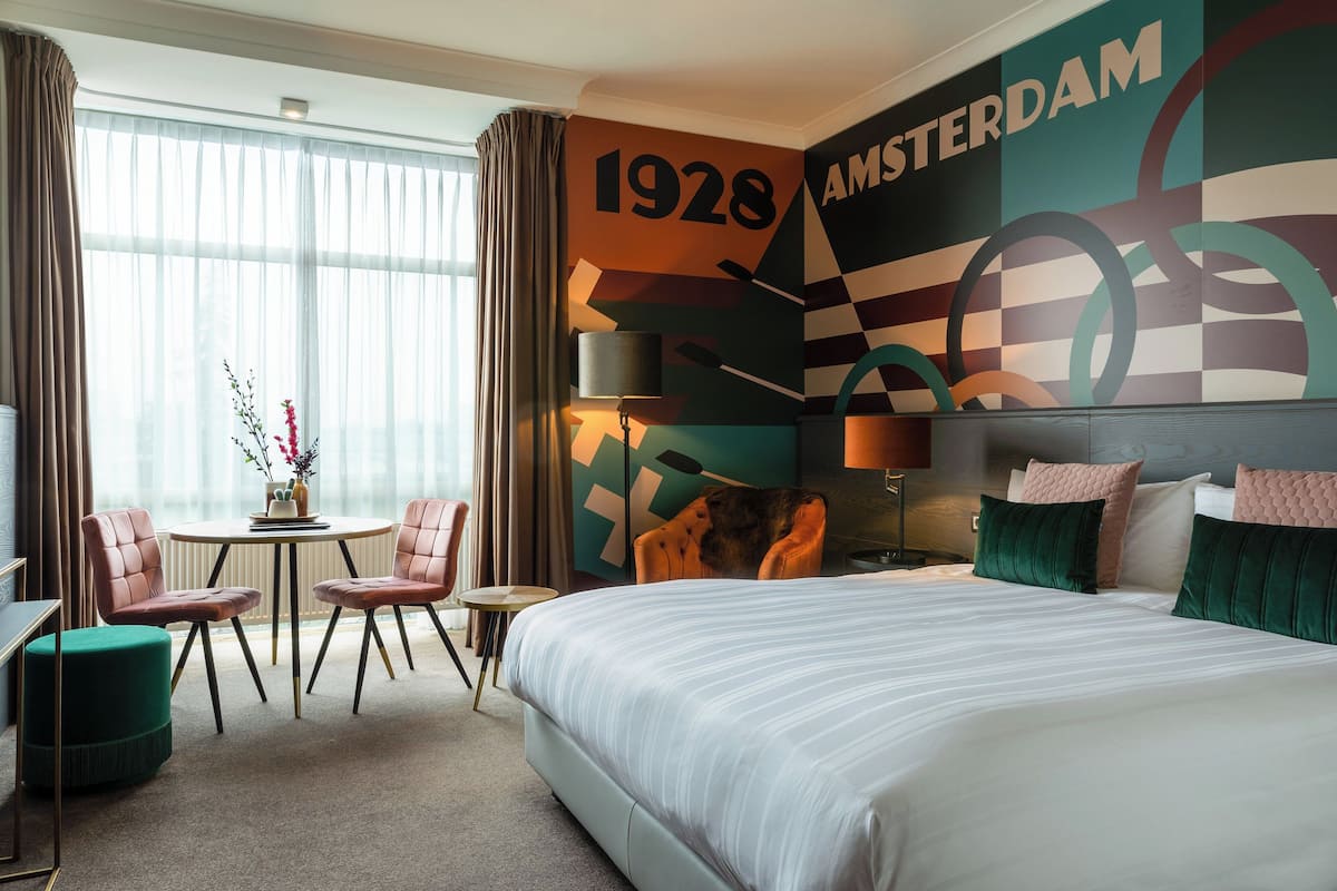 A hotel room with a large bed, a desk with two chairs, a small table with a vase of flowers, and a wall with the year 1928 and the word "AMSTERDAM".