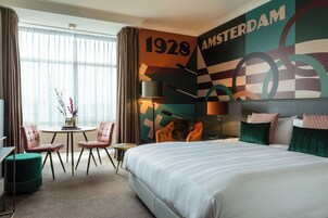 Ein Hotelzimmer mit einem großen Bett, einem Schreibtisch mit zwei Stühlen, einem kleinen Tisch mit Blumen und einer Wand mit dem Jahr 1928 und dem Wort 'AMSTERDAM'.