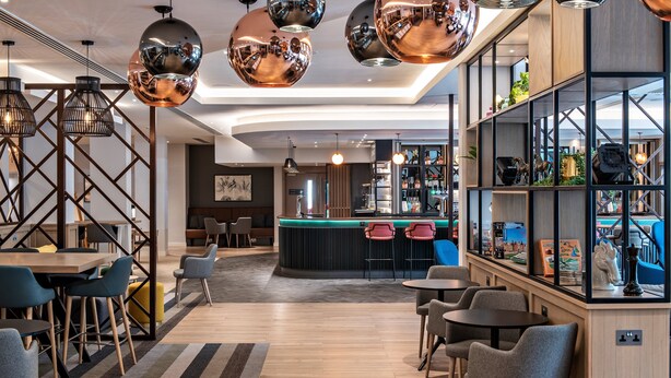 A modern bar area with a counter, seating, and a variety of bottles and decorative items. - Holiday Inn London - Shepperton by IHG (Shepperton)