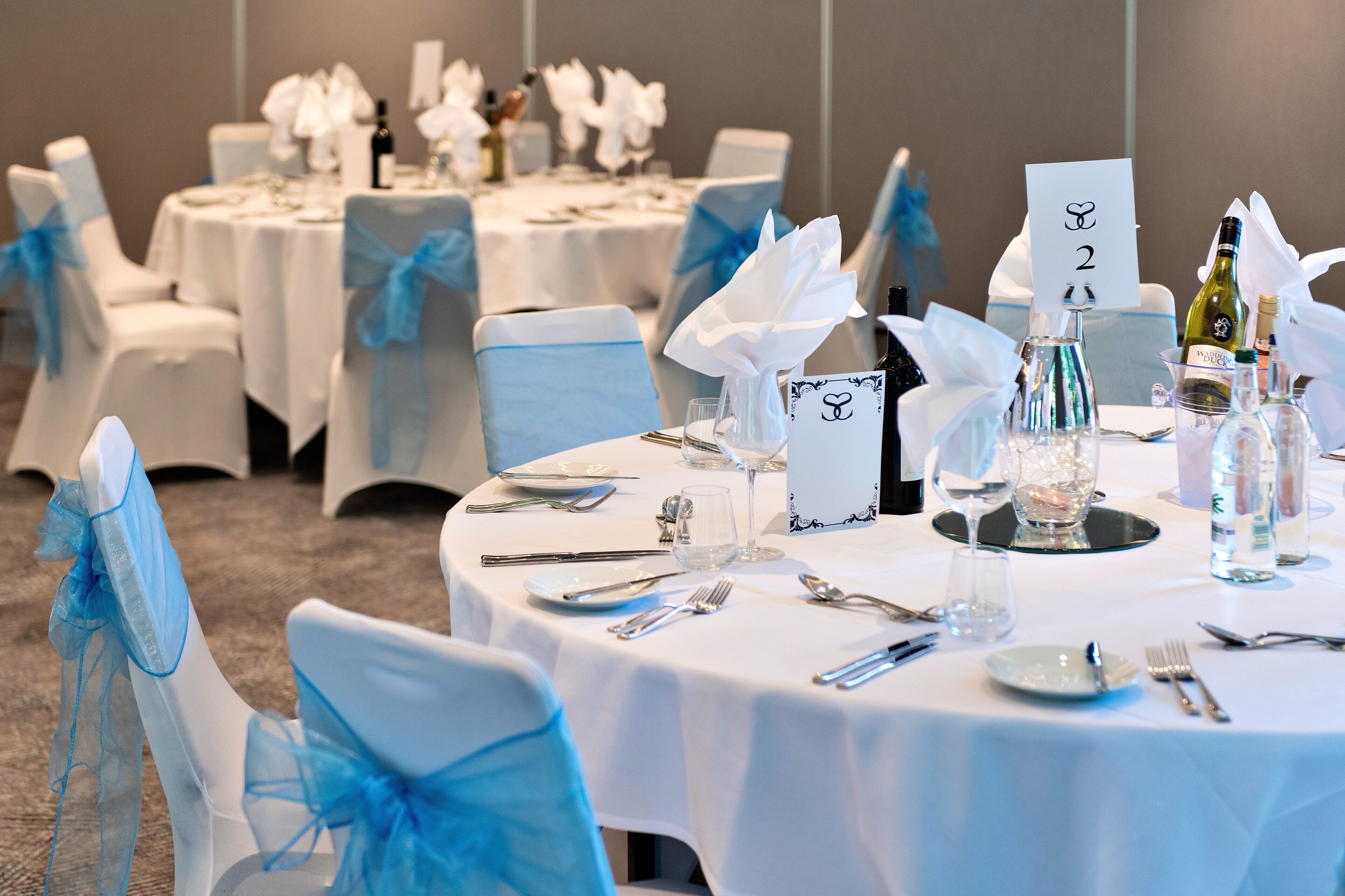 A banquet hall with round tables set for a formal event, each with a number card, white tablecloths, and blue chair sashes.
