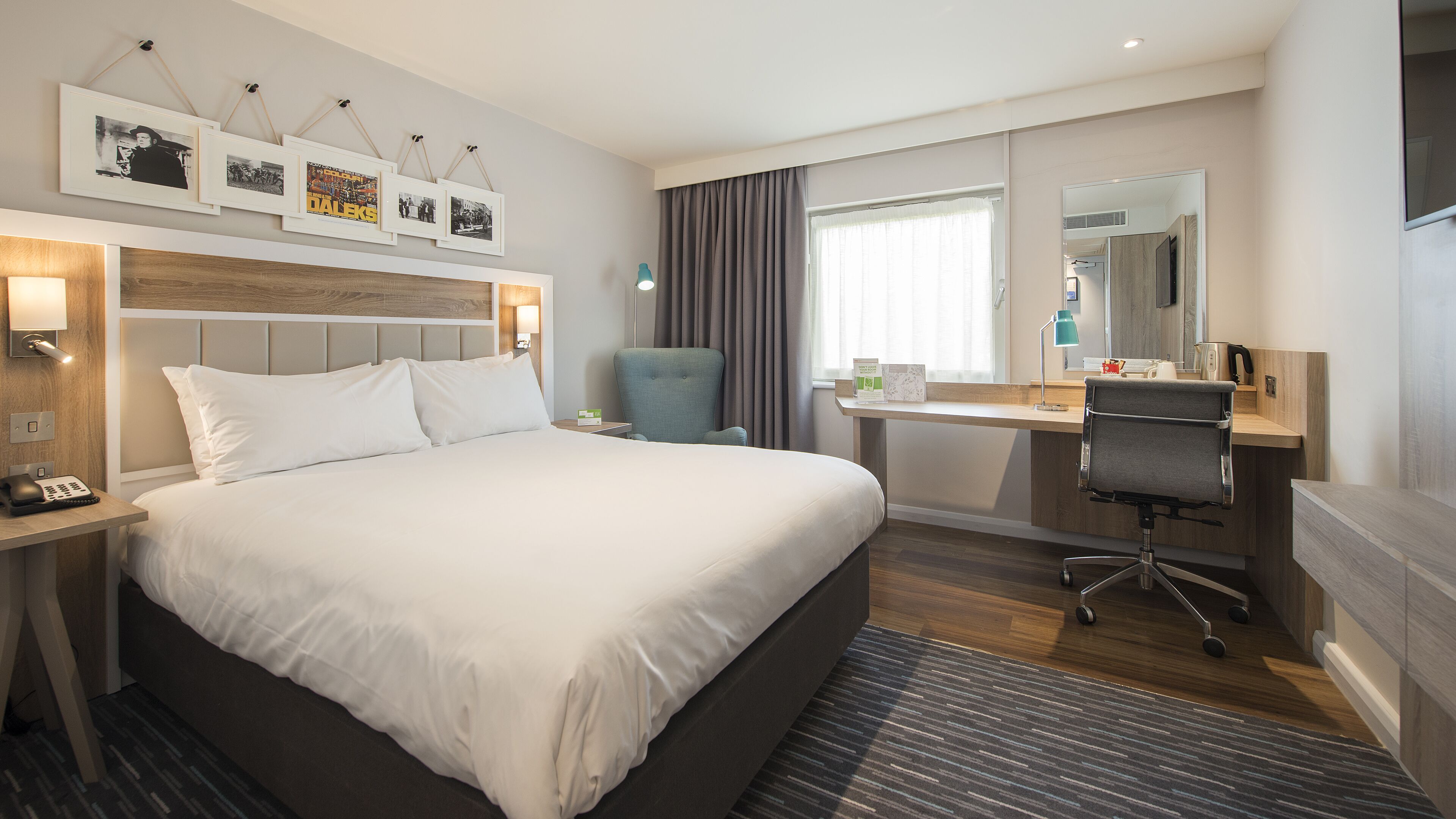 A hotel room with a large bed, a desk with a chair, a small table with a lamp, a mirror, and framed pictures on the wall.