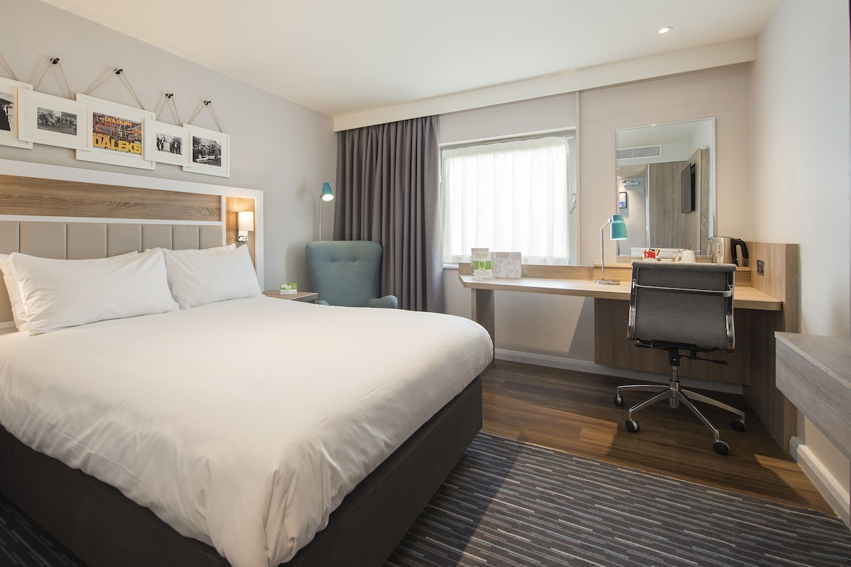 A hotel room with a large bed, a desk with a chair, a small table with a lamp, a mirror, and framed pictures on the wall.