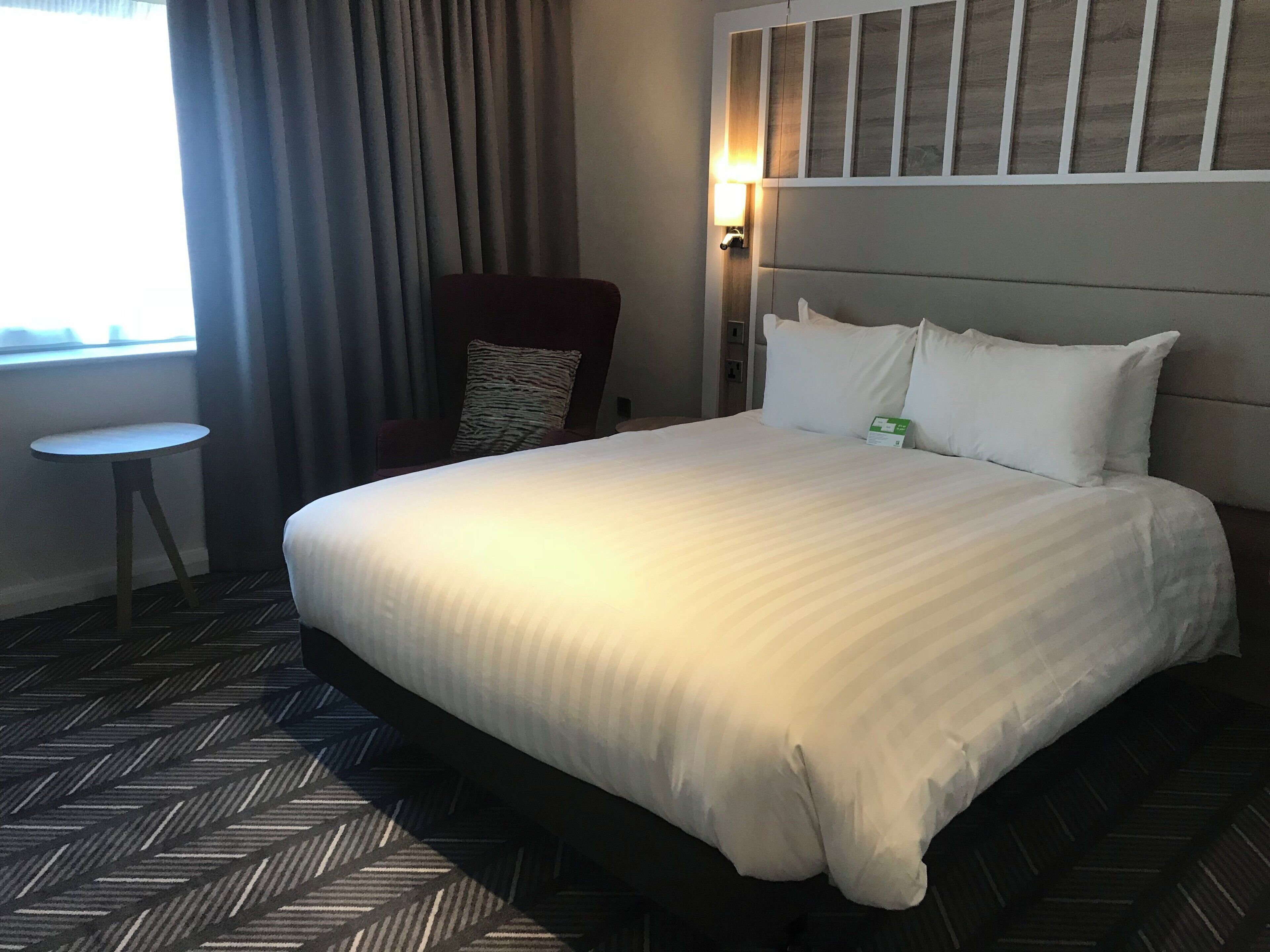 A hotel room with a large bed, a small round table, a chair, and a window with curtains.