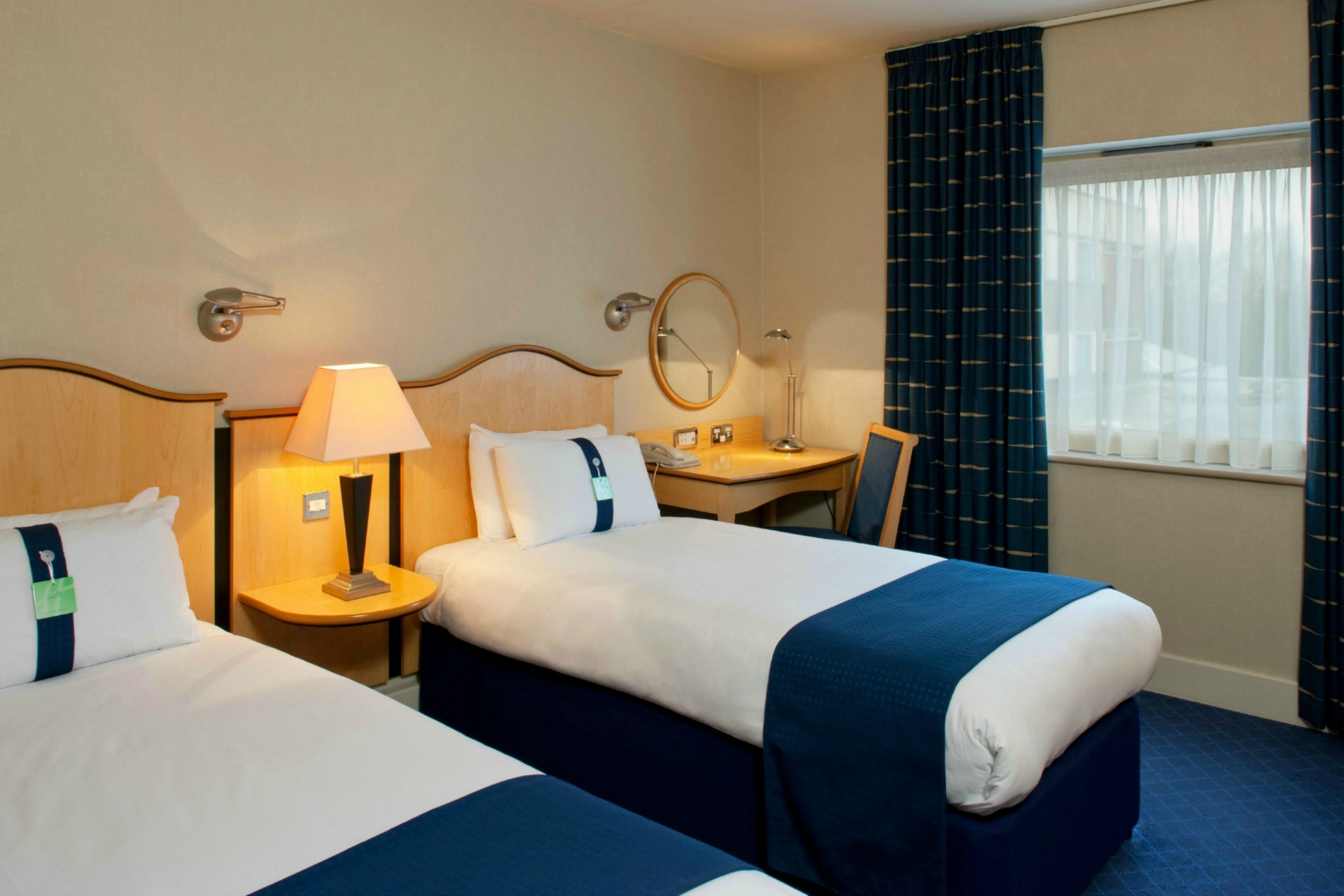 Habitación de hotel con dos camas, un escritorio, una silla, una lámpara, un espejo y una ventana con cortinas.