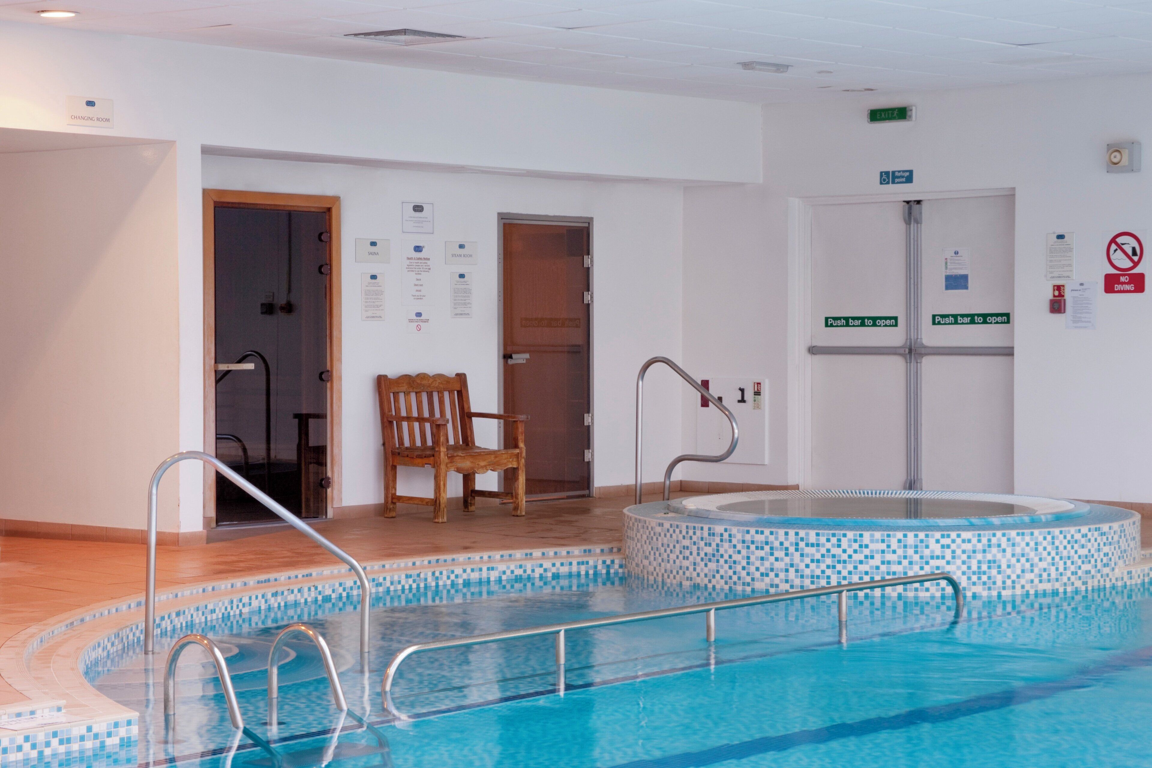 Une piscine avec une chaise en bois et un jacuzzi, le tout entouré de murs blancs portant des panneaux de sécurité.