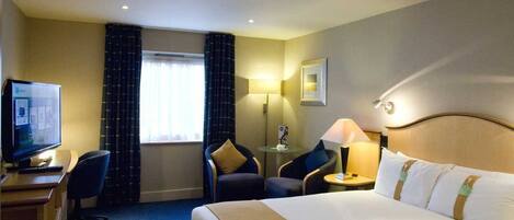 A hotel room with a bed, a desk with a computer, two chairs, a lamp, and a window with curtains.