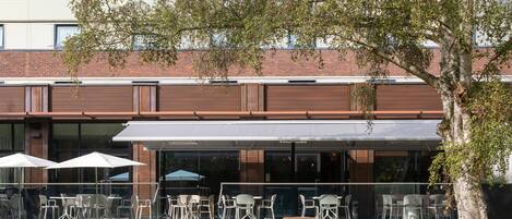 An outdoor seating area with tables and chairs, a covered patio, and a brick building facade.