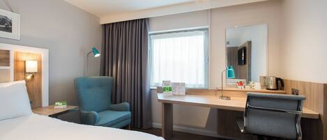 A hotel room with a bed, a desk, a chair, a desk lamp, a window with curtains, a mirror, and a coffee maker.