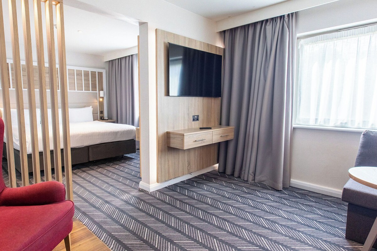 A hotel room with a bed, a desk with a TV, a red chair, and a window with curtains.