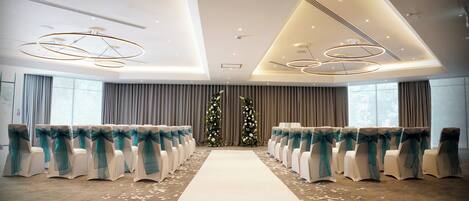 Un salón de bodas con alfombra blanca en el pasillo, filas de sillas adornadas con lazadas turquesas y dos arreglos florales.