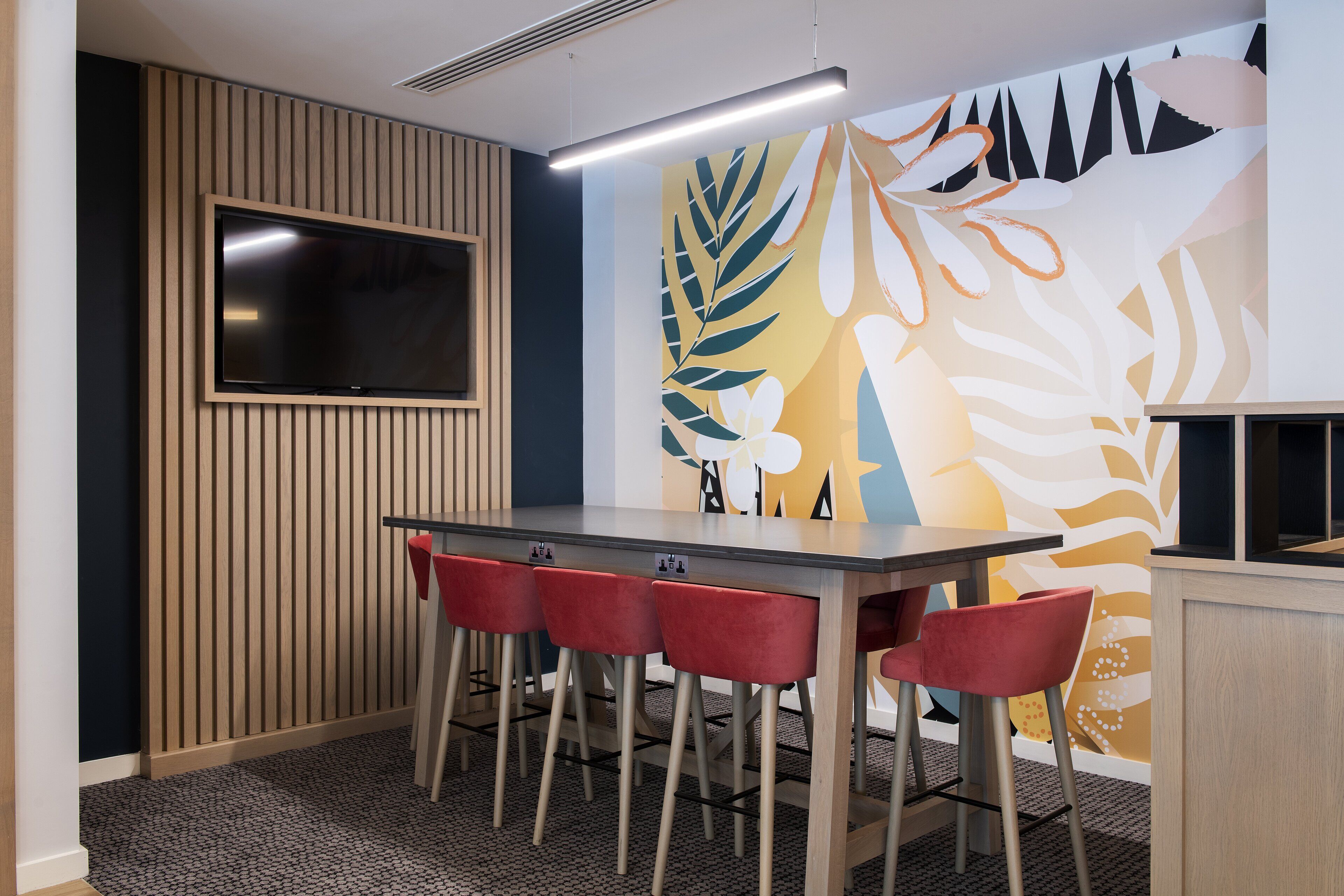 Una sala de conferencias con una gran mesa, cuatro sillas rojas, un televisor de pantalla plana y un mural de pared colorido.