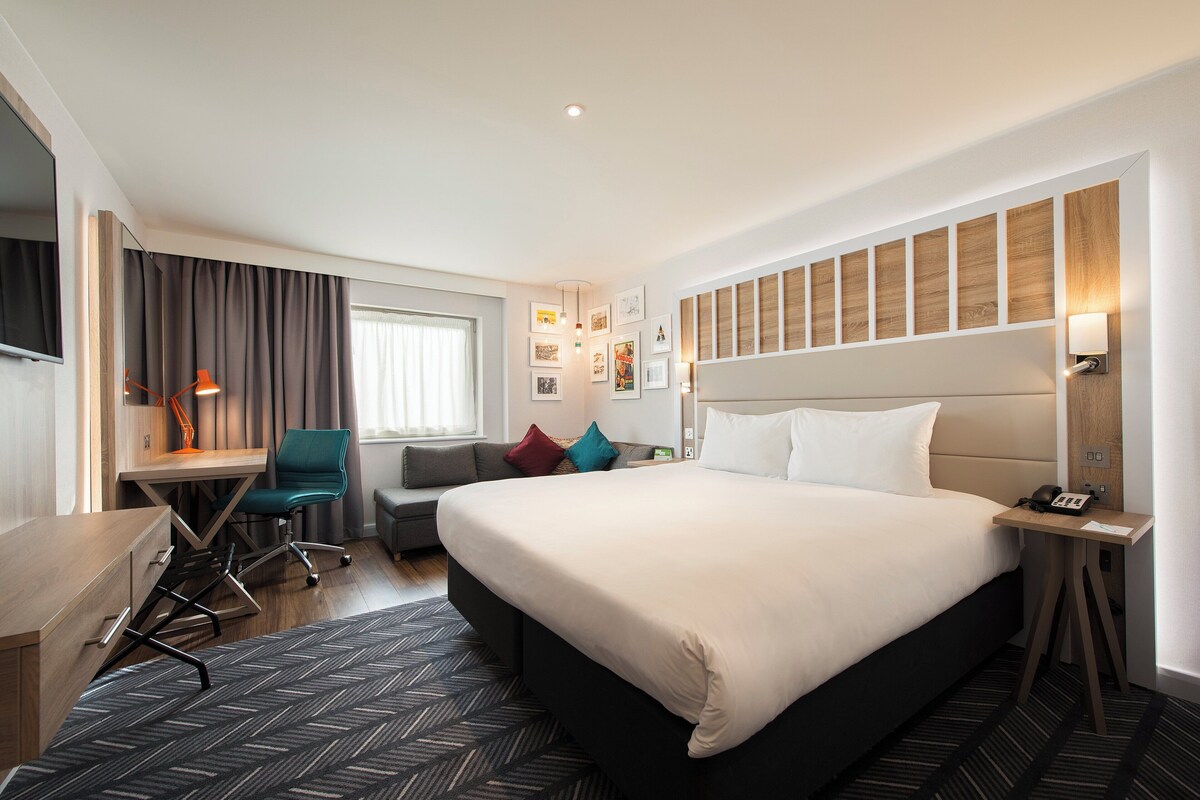 A modern hotel room with a large bed, a desk, a chair, and a sofa.