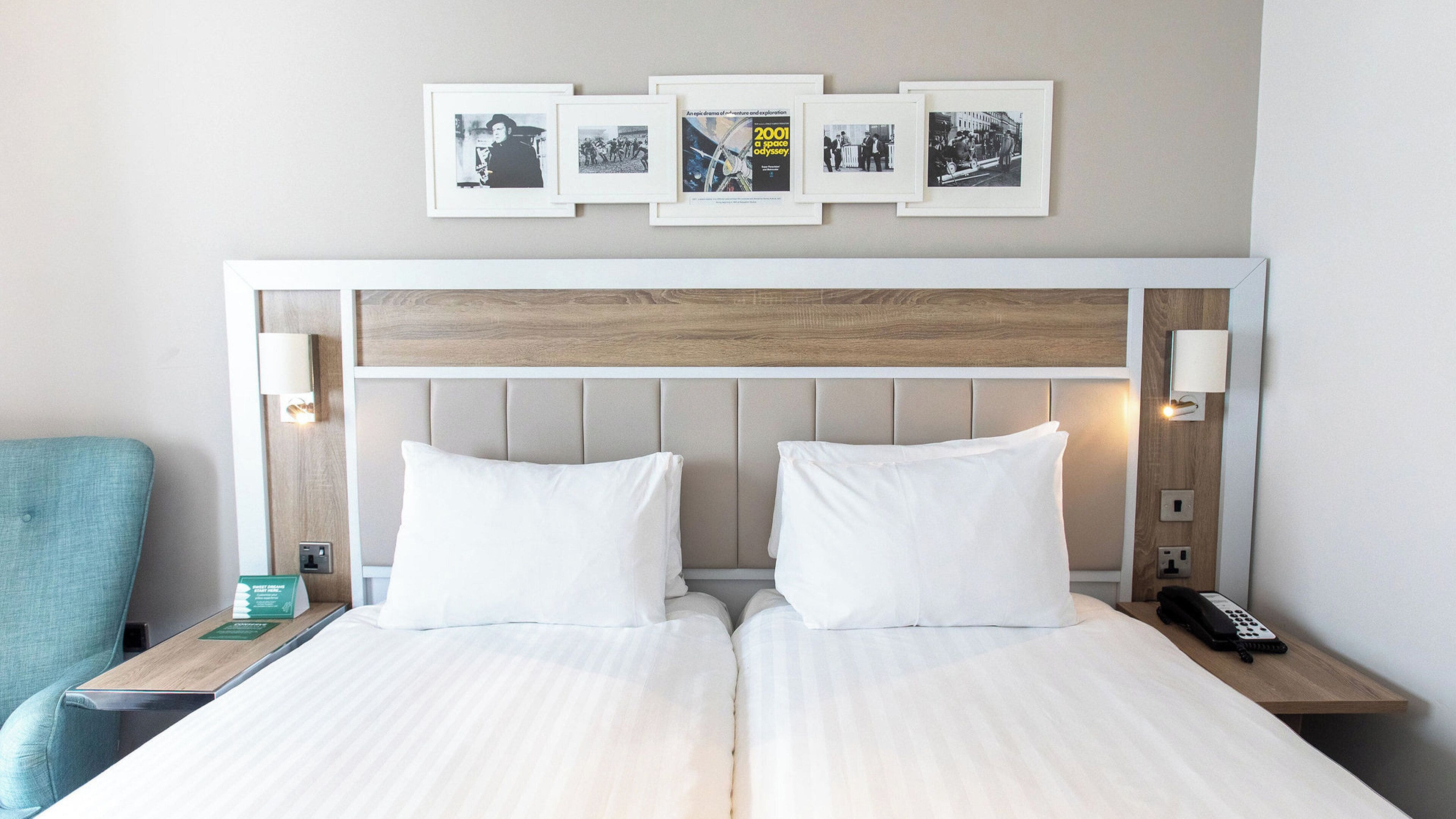 A double bed with white linens, a wooden headboard, bedside lamps, a nightstand with a phone, and framed pictures on the wall.
