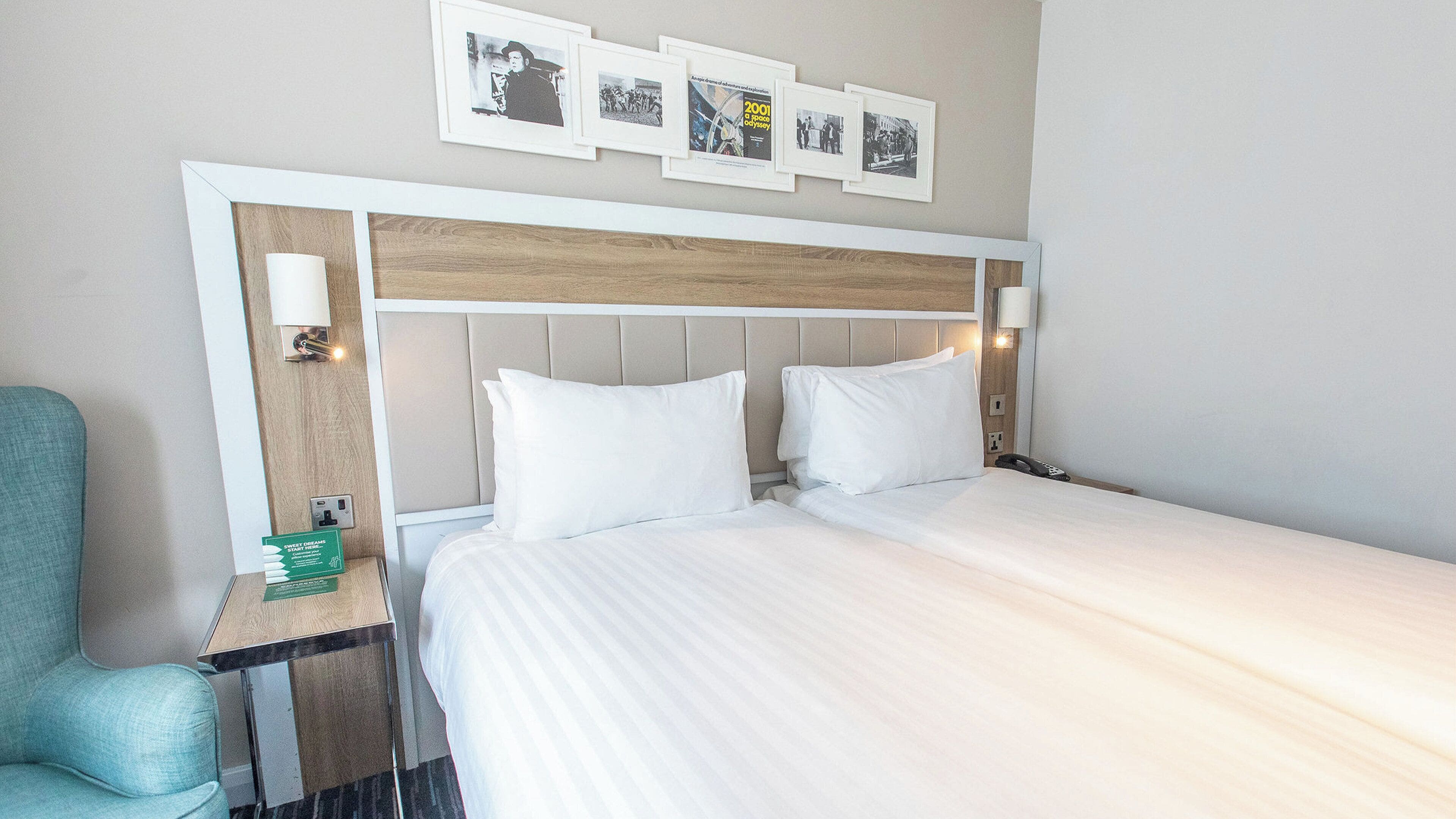 A hotel room with a bed, a nightstand, a chair, and a wall with framed pictures.