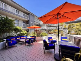 An outdoor patio with blue furniture and orange umbrellas, surrounded by a multi-story building with balconies.