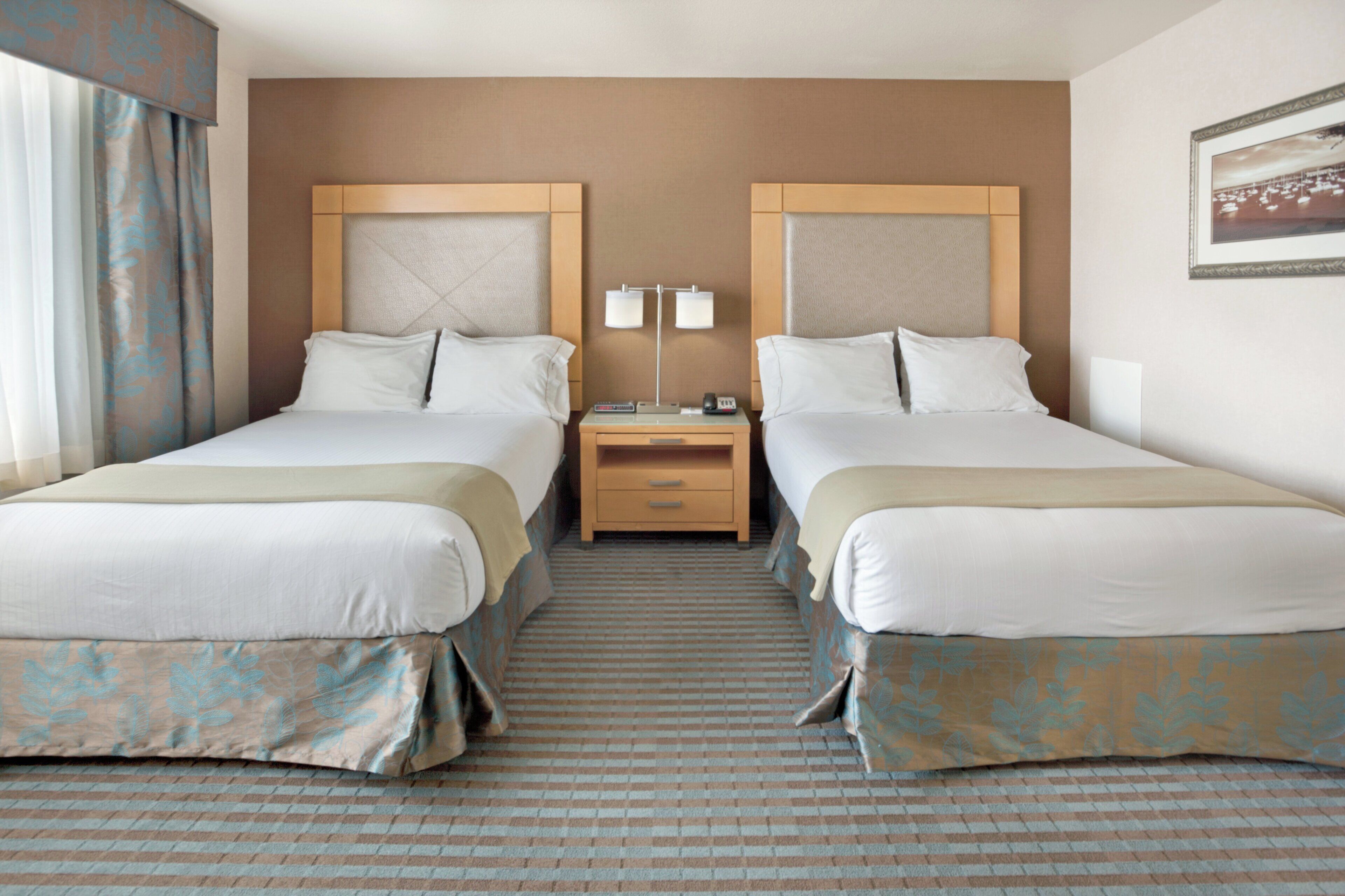 A hotel room with two beds, a nightstand, a lamp, and a framed picture on the wall.