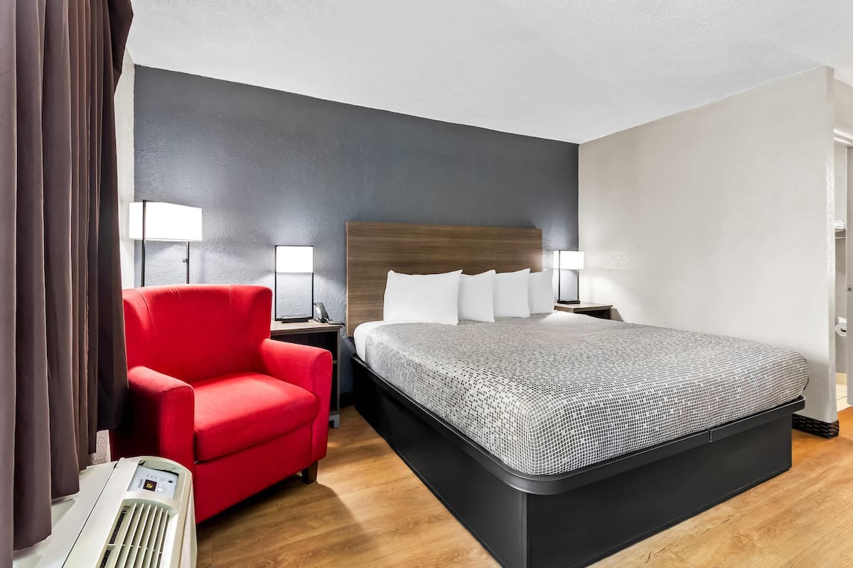A hotel room with a large bed, a red armchair, two bedside lamps, and a wall-mounted heater.