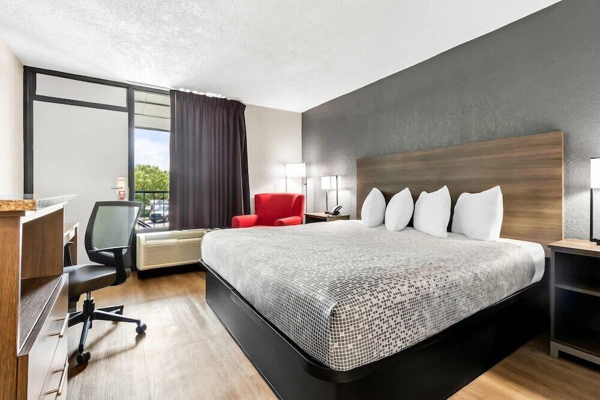 A modern hotel room with a large bed, a desk with a chair, a red armchair, and a view of the outdoors.