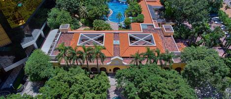 An aerial view of a hotel with a swimming pool, lush greenery, and a distinctive roof design.