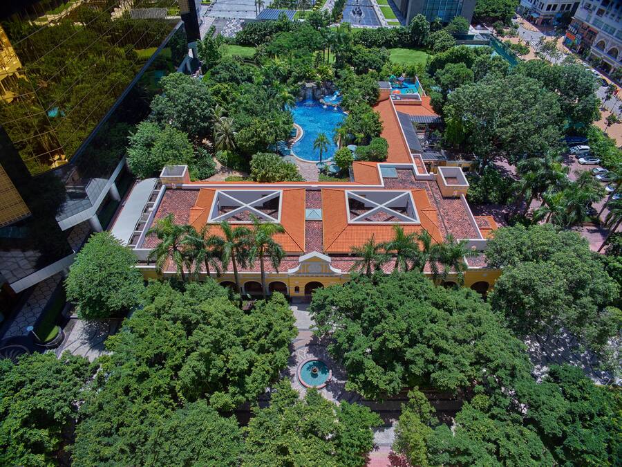 An aerial view of a hotel with a swimming pool, lush greenery, and a distinctive roof design.