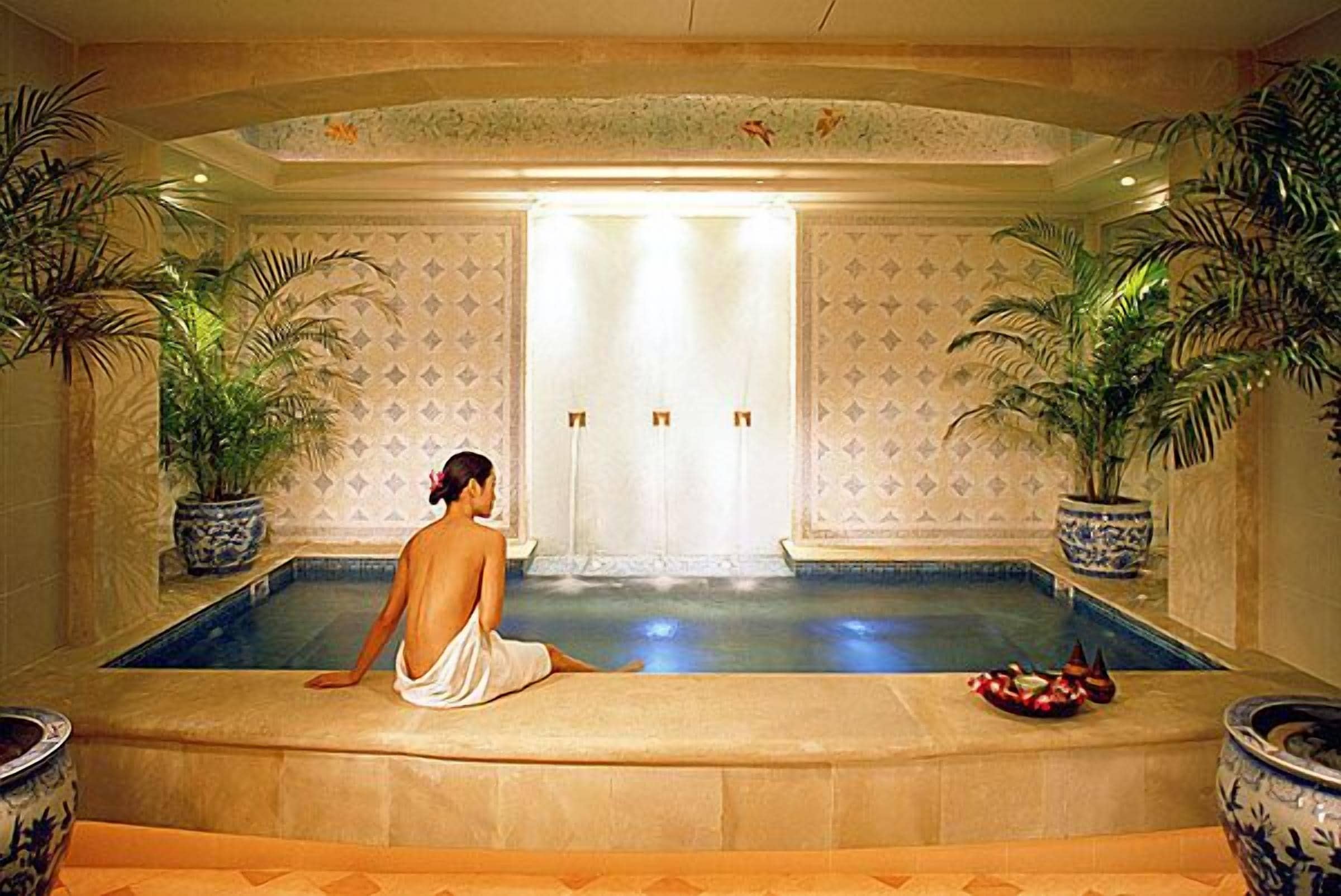 A man sitting by a pool with a decorative wall and potted plants.