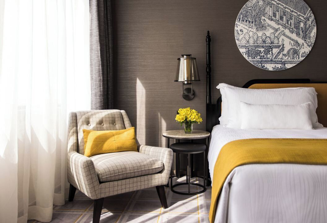 A hotel room with a bed, a chair, a small table with flowers, and a wall-mounted lamp.