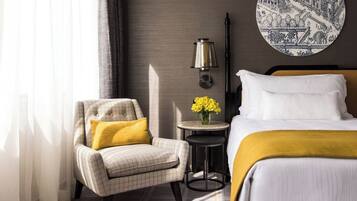 A hotel room with a bed, a chair, a small table with flowers, and a wall-mounted lamp.