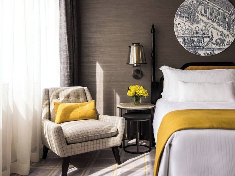A hotel room with a bed, a chair, a small table with flowers, and a wall-mounted lamp.