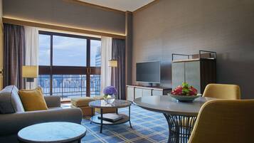Habitación de hotel con sofá, dos sillones, una mesa de centro, televisor y una mesa de comedor con un cuenco de flores.
