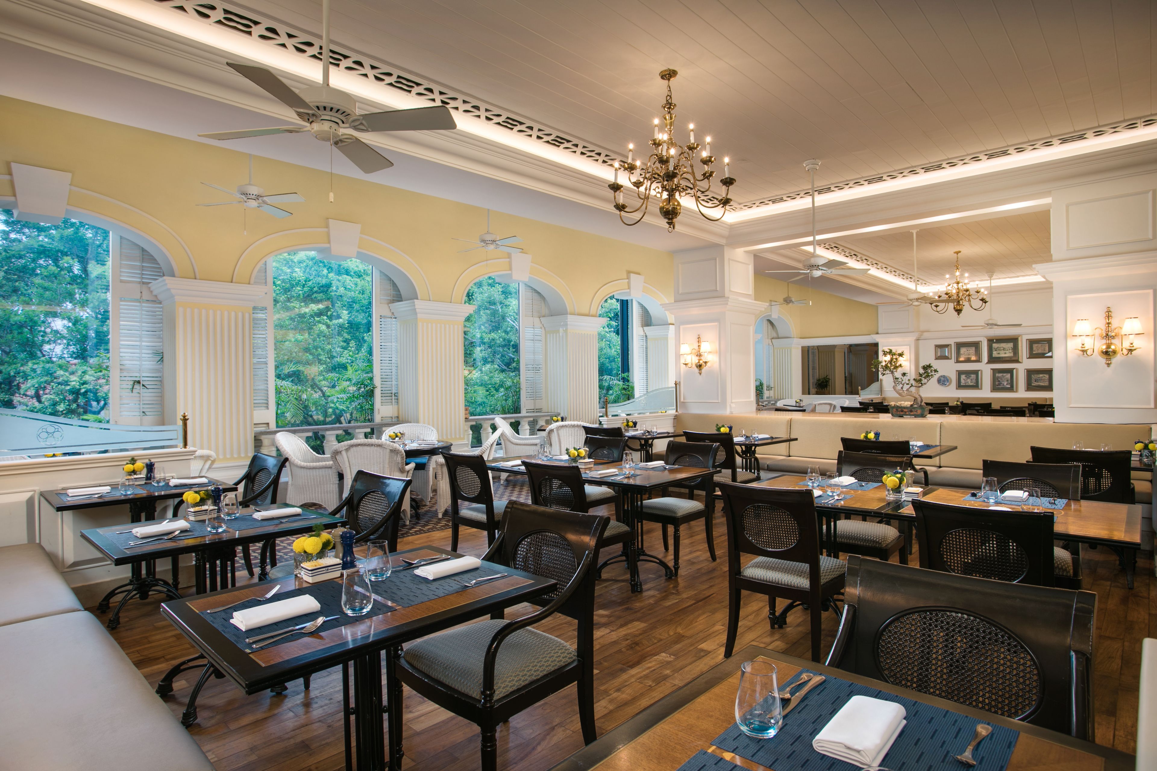 A restaurant with high ceilings, chandeliers, and large windows offering views of greenery. Tables are set with napkins, cutlery, and glasses.