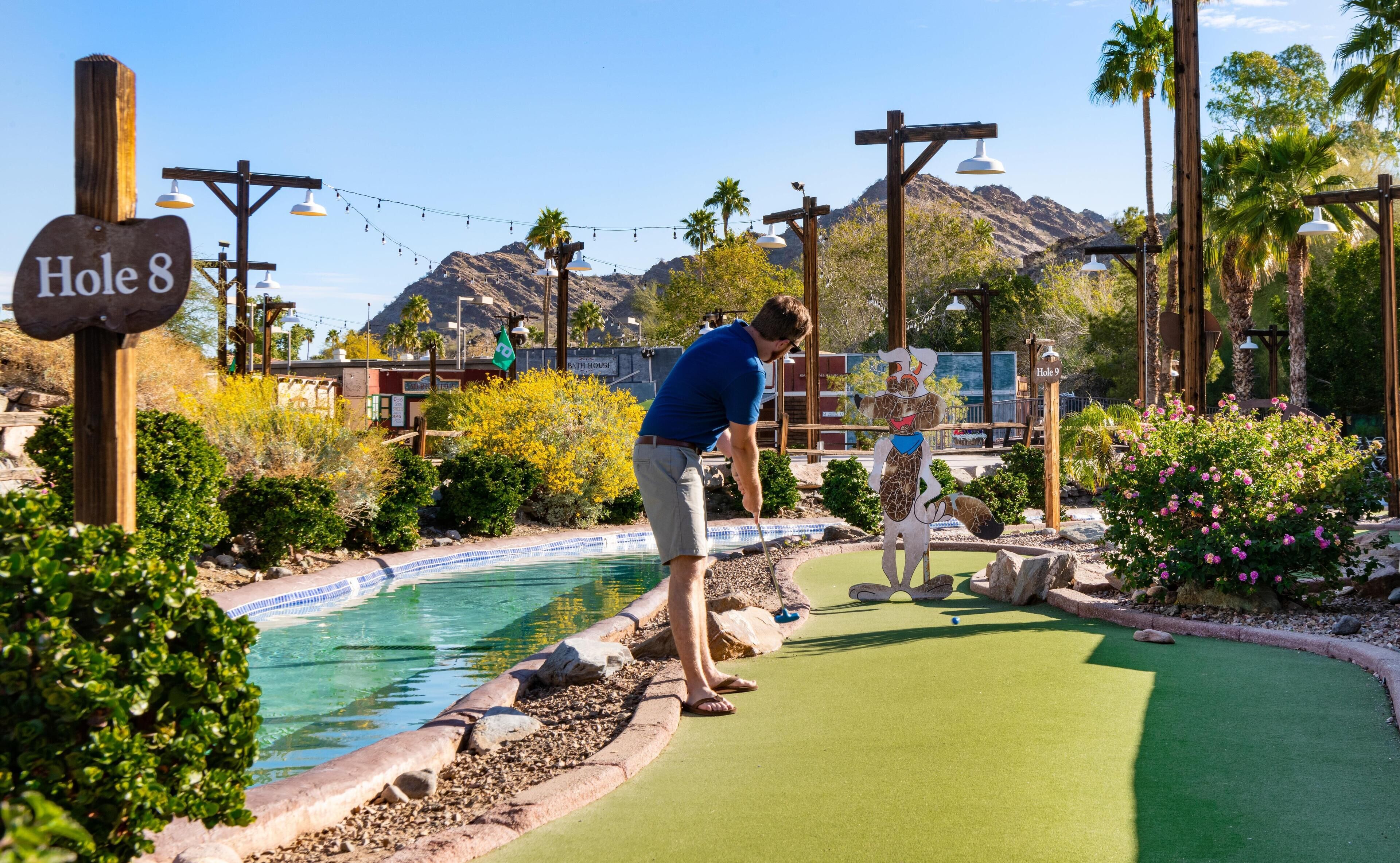 A miniature golf course with a sign for Hole 8, surrounded by greenery and a pool.