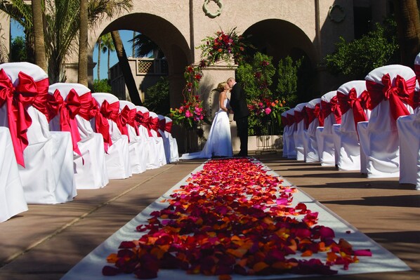 Une allée où se déroule la cérémonie de mariage, avec des chaises blanches et des nœuds rouges, un couple qui s’embrasse et une arche fleurie.