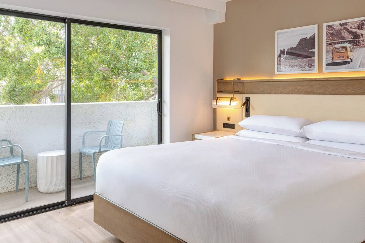 A hotel room with a large bed, a balcony with chairs, and a wall-mounted lamp.