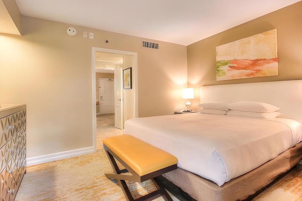 A hotel room with a bed, a bench, a nightstand with a lamp, and a painting on the wall.