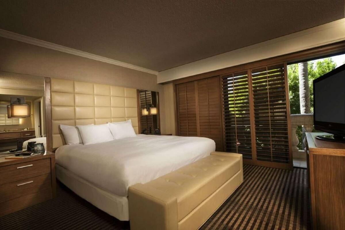 A hotel room with a large bed, a desk with a TV, a bedside table, and a window with blinds.