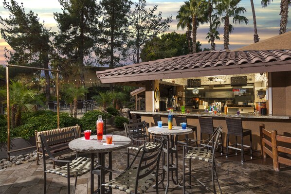 Une terrasse extérieure avec des tables et des chaises rondes, un espace bar et une vue sur des arbres et un bâtiment en arrière-plan.